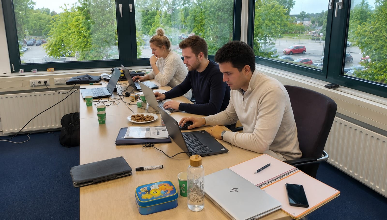Drie cursisten werken aan hun laptops tijdens een praktijkgerichte training bij het raam